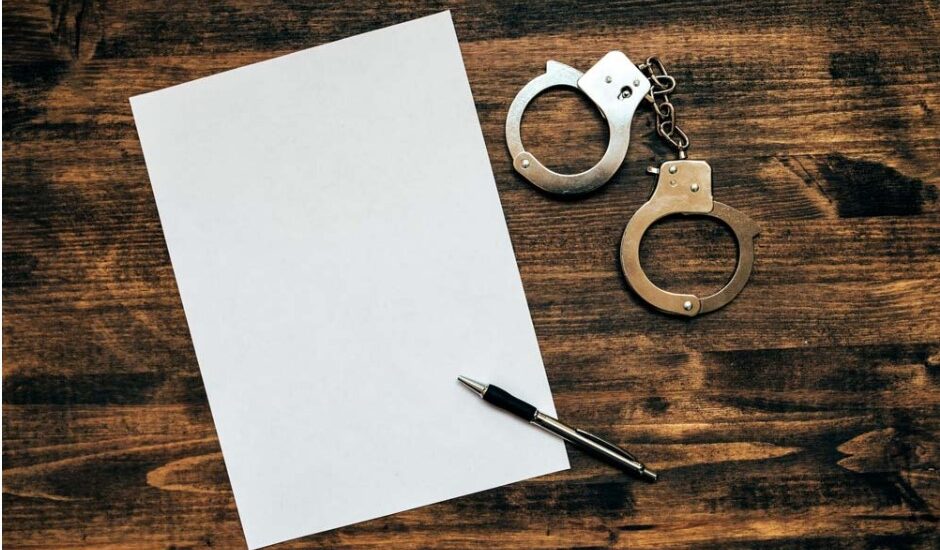 Une feuille blanche, un stylo et une paire de menottes sur un bureau en bois, représentant un rapport de police dans une affaire de conduite avec facultés affaiblies.