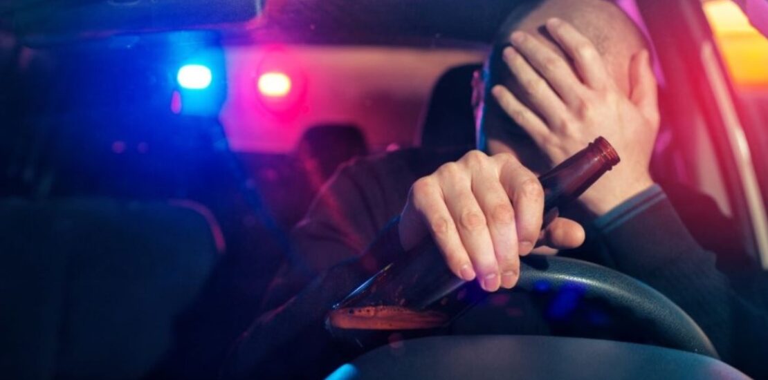 Man holding an empty beer bottle behind the wheel with police lights in the background, depicting a DUI.