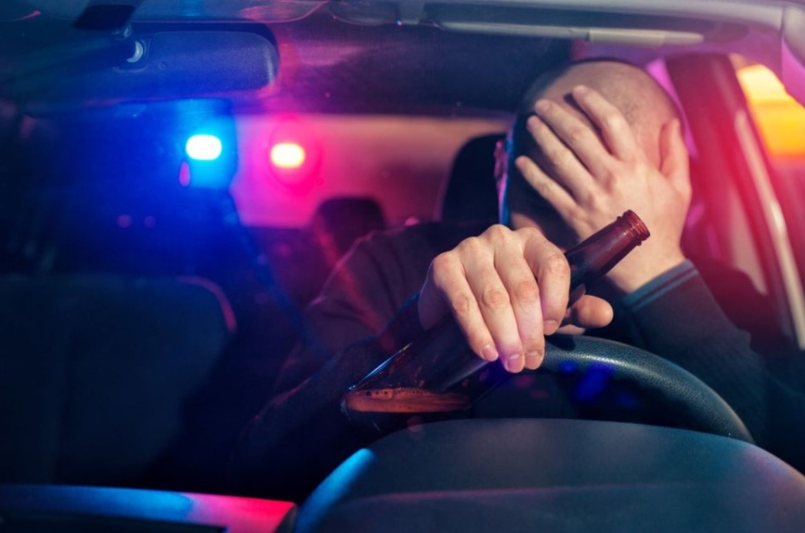 Un homme tenant une bière au volant avec des gyrophares de police en arrière-plan, illustrant l'alcool au volant.