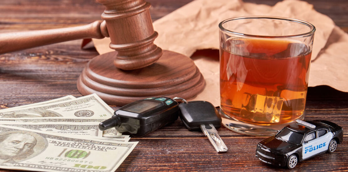 Money, car keys, a toy police car, and a glass of alcohol on a table with a judge's gavel, representing the risks of impaired driving.