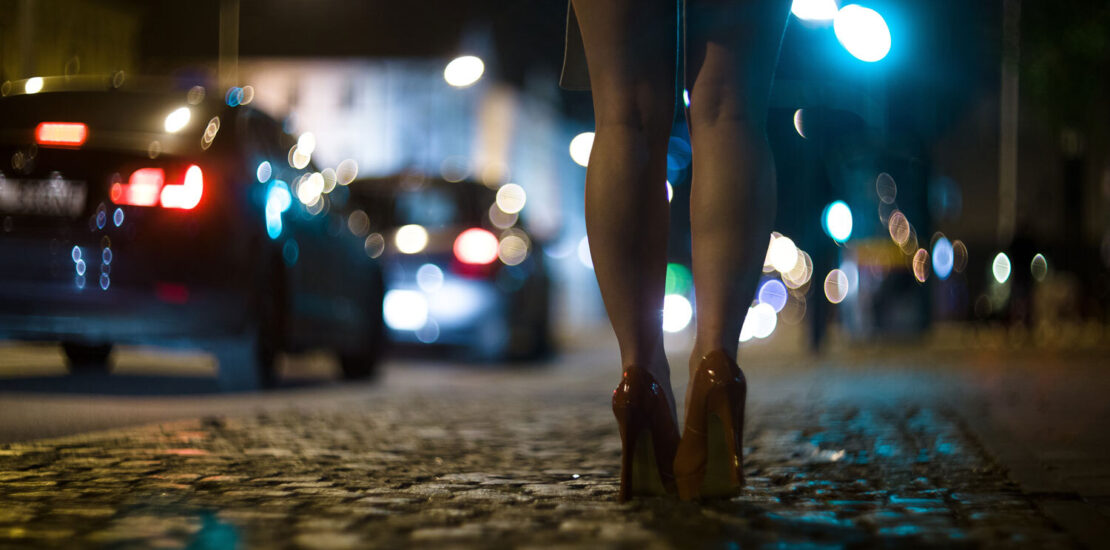 Une femme debout en talons rouges sur le trottoir avec des voitures qui passent, suggérant le besoin d'un avocat criminel pour des activités sexuelles.